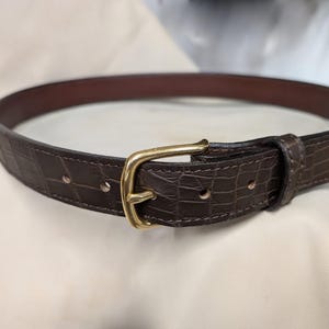 Op de afbeelding: Een donkerbruine riem met een gestructureerd krokodil-reli&euml;fpatroon op de band. De riem wordt vastgezet met een gepolijste goudkleurige metalen gesp en is voorzien van een bijpassende lederen lus. De binnenkant van de band is glad donkerbruin.