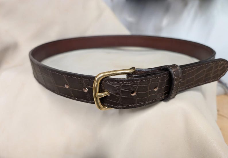 Op de afbeelding: Een donkerbruine riem met een gestructureerd krokodil-reli&euml;fpatroon op de band. De riem wordt vastgezet met een gepolijste goudkleurige metalen gesp en is voorzien van een bijpassende lederen lus. De binnenkant van de band is glad donkerbruin.