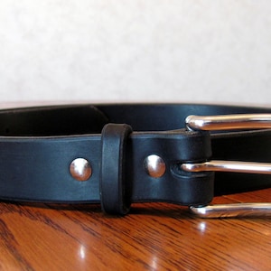 May include: A black leather belt with a silver buckle and two silver rivets. The belt is on a wooden surface.