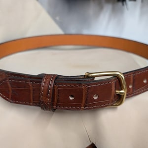 Op de afbeelding: Een bruine leren riem met een krokodil-reli&euml;fpatroon, voorzien van een glanzende goudkleurige metalen gesp, een bijpassende leren lus en een gladde, lichtbruine binnenvoering met zichtbare stiksels langs de randen.