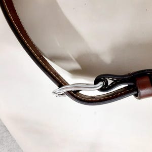 May include: Close-up of a brown leather strap with white stitching and a silver-colored metal clasp. The strap is curved and appears to be part of a larger item, possibly a bag or belt.