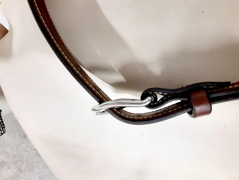 May include: Close-up of a brown leather strap with white stitching and a silver-colored metal clasp. The strap is curved and appears to be part of a larger item, possibly a bag or belt.