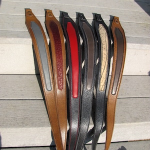 May include: Seven leather guitar straps in various colors, including brown, gray, red, black, and beige. The straps have a contrasting leather strip down the center.
