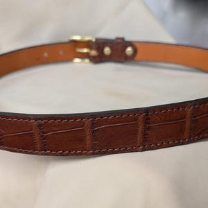 Op de afbeelding: Een bruine riem met een gestructureerd krokodil-reli&euml;fpatroon aan de buitenkant en een gladde, lichtbruine voering aan de binnenkant. De riem is voorzien van een gepolijste goudkleurige metalen gesp en bijpassende goudkleurige klinknagels.