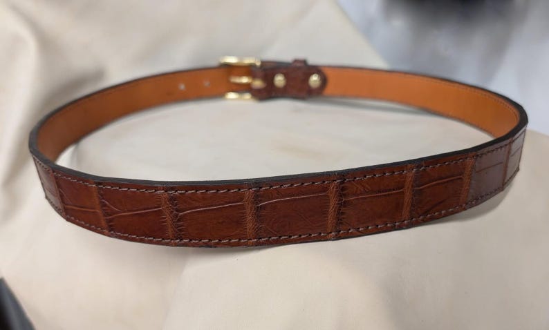 Op de afbeelding: Een bruine riem met een gestructureerd krokodil-reli&euml;fpatroon aan de buitenkant en een gladde, lichtbruine voering aan de binnenkant. De riem is voorzien van een gepolijste goudkleurige metalen gesp en bijpassende goudkleurige klinknagels.