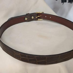 Op de afbeelding: Een donkerbruine riem met een gestructureerd krokodil-reli&euml;fpatroon op de buitenkant. De riem is voorzien van een gladde bruine leren voering aan de binnenzijde, ton-sur-ton stiksels langs de randen en een goudkleurige metalen gesp met twee pinnen.