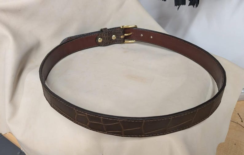 Op de afbeelding: Een donkerbruine riem met een gestructureerd krokodil-reli&euml;fpatroon op de buitenkant. De riem is voorzien van een gladde bruine leren voering aan de binnenzijde, ton-sur-ton stiksels langs de randen en een goudkleurige metalen gesp met twee pinnen.