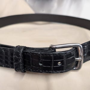 Op de afbeelding: Een zwarte riem met een gestructureerd krokodillenpatroon over de gehele band. De riem is voorzien van een gepolijste zilverkleurige metalen gesp, een bijpassende lus en een puntig uiteinde, afgewerkt met zichtbare stiksels.