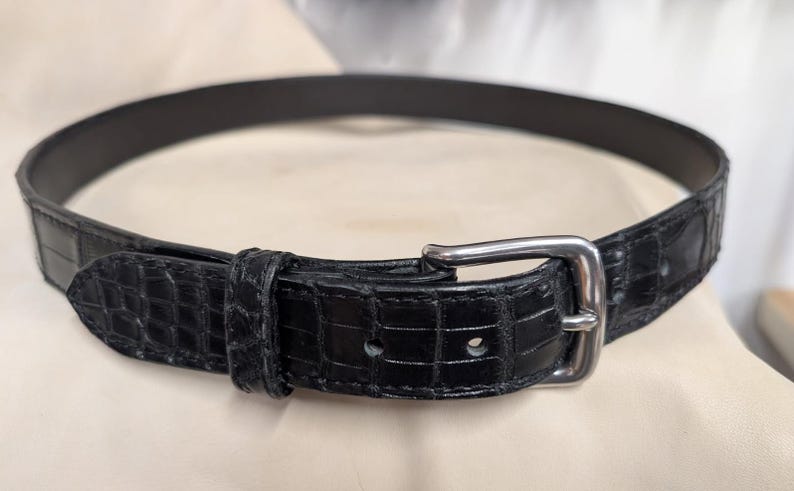 Op de afbeelding: Een zwarte riem met een gestructureerd krokodillenpatroon over de gehele band. De riem is voorzien van een gepolijste zilverkleurige metalen gesp, een bijpassende lus en een puntig uiteinde, afgewerkt met zichtbare stiksels.