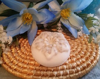 May include: A round, white soap with a floral design sits on a woven gold-colored mat. The soap is surrounded by blue and white flowers. The soap has a light brown speckled pattern.