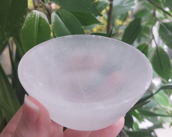 Puede incluir: Un pequeño cuenco de selenita blanco translúcido con forma redondeada. El cuenco se sostiene en una mano, con follaje verde en el fondo. El cuenco se utiliza probablemente para guardar cristales u otros objetos pequeños.