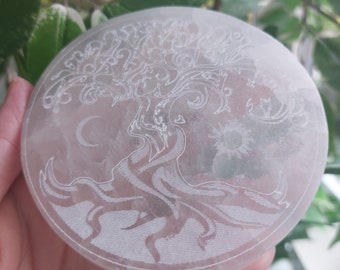 Peut inclure: Disque en cristal de sélénite blanc avec un motif d'arbre de vie gravé. L'arbre a des branches et des racines complexes, et une lune croissante est visible en arrière-plan.