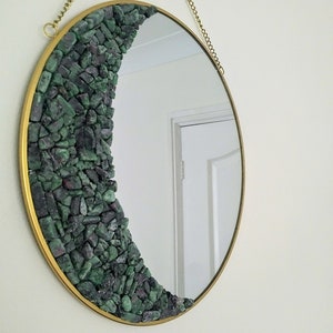 May include: A round mirror with a gold frame and a half-circle design of green and grey stones. The mirror is hanging from a gold chain.