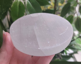 May include: A round, white selenite crystal charging plate. The plate is smooth and has a slightly translucent appearance. The background features green leaves.