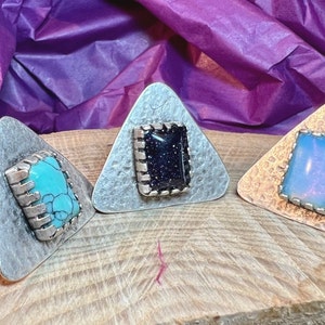 May include: Three silver triangle rings with different colored stones. The rings have a textured surface and a square stone set in the center. The stones are turquoise, black, and opal.