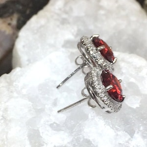 May include: A pair of silver earrings with red oval-shaped gemstones surrounded by a halo of smaller clear gemstones.