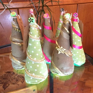 May include: Five miniature Christmas trees made of fabric with polka dots and burlap. The trees are decorated with gold ribbon, gold bells, and a star on top. The trees are sitting on a glass table.