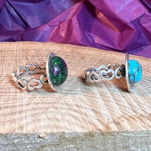 Op de afbeelding: Twee zilveren ringen met hartvormige ontwerpen. De ring aan de linkerkant heeft een groene en roze edelsteen, terwijl de ring aan de rechterkant een blauwe en bruine edelsteen heeft.