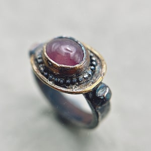 May include: A silver ring with a large, oval, purplish-pink gemstone. The ring has gold-coloured accents around the stone and small, dark embellishments. The design is detailed, with a vintage or artisan style. The ring is a piece of jewellery.