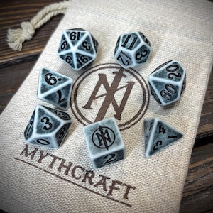 May include: A collection of seven dice in different shapes, featuring white numerals on a grey surface, displayed on a beige cloth bag. The bag is printed with the text "MYTHCRAFT" and a logo. These dice are for tabletop games.
