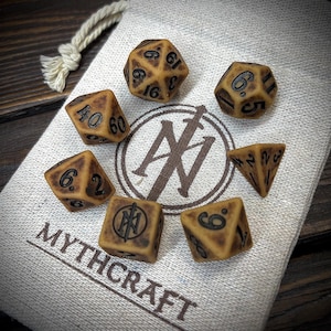 May include: A set of seven tan and black dice for role-playing games, displayed on a canvas bag. The dice are in various shapes, including a 20-sided die, a 12-sided die, and a 4-sided die. The bag has the text "MYTHCRAFT" printed on it.