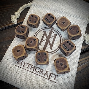 May include: Ten brown dice with black pips are displayed on a beige drawstring bag. The dice have a worn, aged look. The bag has a logo and the text "MYTHCRAFT".