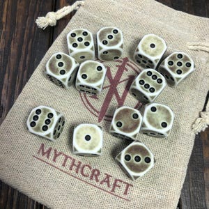 Puede incluir: Un conjunto de diez dados de hueso en una bolsa de cordón beige. Los dados son blancos con puntos negros y tienen un aspecto moteado y envejecido. La bolsa tiene la palabra "MYTHCRAFT" impresa en rojo.