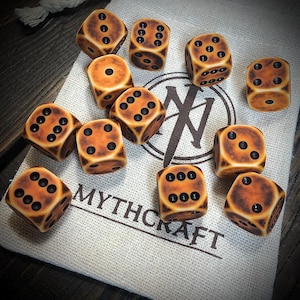 Può includere: Un set di dodici dadi dall'aspetto invecchiato e usurato. I dadi sono beige con bordi marrone scuro e punti neri. Sono disposti su un sacchetto di tela con la scritta "MYTHCRAFT" stampata.