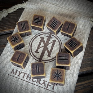 Può includere: Un set di dieci pietre runiche marroni e beige con simboli incisi, presentate su un sacchetto di stoffa beige. La borsa ha un logo circolare con la scritta "MYTHCRAFT". Le rune sono disposte in cerchio.