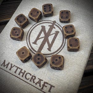 May include: Ten brown dice with black dots are arranged on a tan fabric surface. The dice surround a circular logo with the text "Mythcraft" at the bottom. The logo features a sword and the letters "N" and "I".