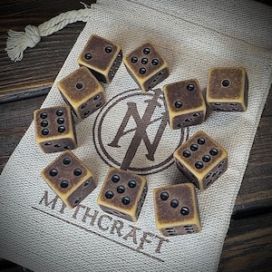Könnte beinhalten: Zehn braune Würfel mit schwarzen Augen liegen auf einem beigen Stoffbeutel. Die Würfel haben ein Vintage-Aussehen. Der Beutel hat ein Logo und den Schriftzug "MYTHCRAFT". Geeignet für Tischspiele und Rollenspiele.