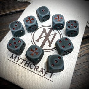 Op de afbeelding: Een set van tien donkergrijze dobbelstenen met rode ogen en een rood kruisontwerp op sommige vlakken. De dobbelstenen liggen op een beige stoffen zak met de tekst "MYTHCRAFT" en een logo. De dobbelstenen zijn voor tabletop games.
