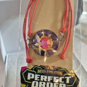 May include: A bracelet featuring a circular pendant with a purple and gold design and a central pink gem, attached to a red cord. The packaging includes a label with the text MEGA EVOLUTION PERFECT ORDER in white, yellow, and black lettering.