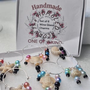 May include: Set of six handmade wine glass charms. Each charm features a silver hoop with decorative beads and a seashell, starfish, or turtle charm. The box reads "Handmade Humbled Acorn Creations".