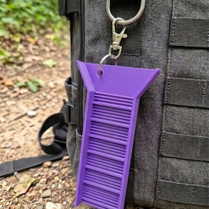 May include: A purple plastic sluice box keychain featuring a flared top and a series of horizontal riffles along its length. The object is attached to a silver-toned metal lobster claw clasp and a split ring, hanging from a dark gray fabric backpack.