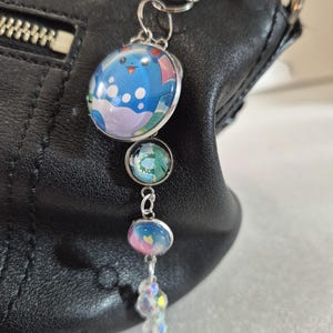 May include: A silver-toned metal keychain featuring a large circular charm with a blue cartoon character graphic, followed by two smaller circular charms with abstract blue and pink patterns, and a dangling clear iridescent crystal bead accent.
