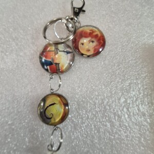 May include: A silver-toned metal keychain featuring a lobster clasp and a split ring. Attached are three circular glass cabochon charms: one depicting a person with red hair, one showing a person holding a bow, and one with a swirling yellow pattern.