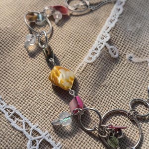 May include: A delicate necklace featuring a series of silver-toned rings and a variety of colorful beads. The beads include iridescent, pink, yellow, and green elements. The necklace is displayed on a textured, neutral-toned surface.
