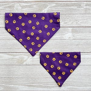 May include: Two purple dog bandanas with a gold paw print pattern. The bandanas are on a light wood background.