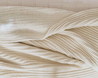 Könnte beinhalten: Eine cremefarbene gestrickte Wolldecke mit Rippenstruktur. Die Decke ist gefaltet und über eine Oberfläche gelegt.