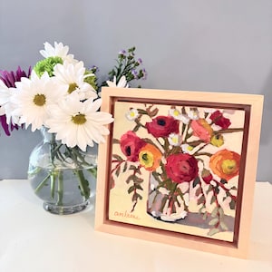 Puede incluir: Pintura floral enmarcada que presenta un ramo de flores rojas, naranjas y blancas en un jarrón transparente. La pintura tiene un fondo beige claro y un marco de madera clara. Un jarrón de margaritas blancas es visible en el fondo.