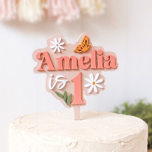 May include: A pink and white cake topper with the name Amelia and the number 1, decorated with flowers and a butterfly. The topper is on a white cake.