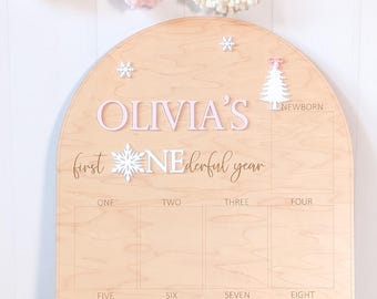 May include: A wooden milestone board with an arched top. The board is light brown with the words "OLIVIA'S first wonderful year" in pink and white. It has spaces for monthly photos and decorative snowflake and tree accents.