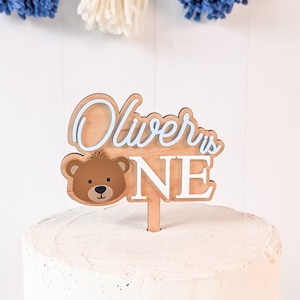 May include: A wooden cake topper with the text "Oliver is ONE" in a layered design. The topper features a brown teddy bear face and is placed on top of a white frosted cake. Decorative yarn tassels hang in the background.