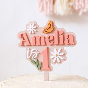 May include: A pink cake topper with the name Amelia and the number 1, surrounded by white flowers and a butterfly. The topper is on a white cake.