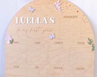 May include: Wooden milestone board with an arched top. The board is light brown with the text "LUELLA'S fairy first year". It has spaces for months one through twelve, plus "Newborn". Adorned with butterflies, a fairy, and a mushroom.