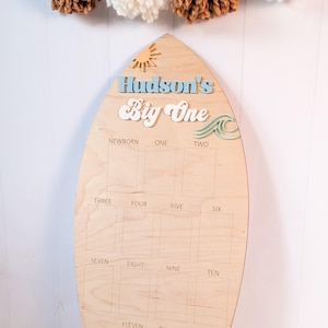Peut inclure: Un tableau de jalons en forme de planche de surf en bois avec le texte "Hudson's Big One". Le tableau comporte des espaces pour les photos de la naissance à douze mois. Éléments décoratifs : soleil, vague et palmier.
