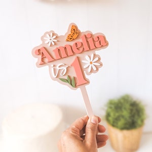 May include: Pink and white cake topper with the name Amelia and the number 1, decorated with flowers and a butterfly. The topper says "Amelia is 1".