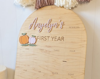 Puede incluir: Un tablero de hitos de madera en forma de arco con el texto grabado Anyelyn's FIRST YEAR. Incluye una sección para NEWBORN y doce cajas rectangulares etiquetadas de ONE a TWELVE, decoradas con dos pequeñas calabazas de color naranja y rosa.