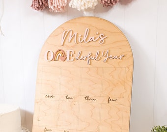 May include: A wooden arch-shaped board with the text "Mila's One-derful Year" and a rainbow graphic. The board is divided into twelve squares, each labeled with a number from one to twelve.